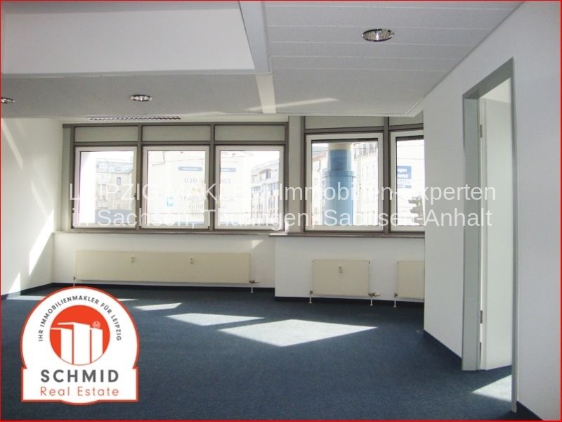 Leerer Büroraum mit Fensterfront, Deckenleuchten, dunklem Bodenbelag, Heizkörpern und Tür zu Nebenraum